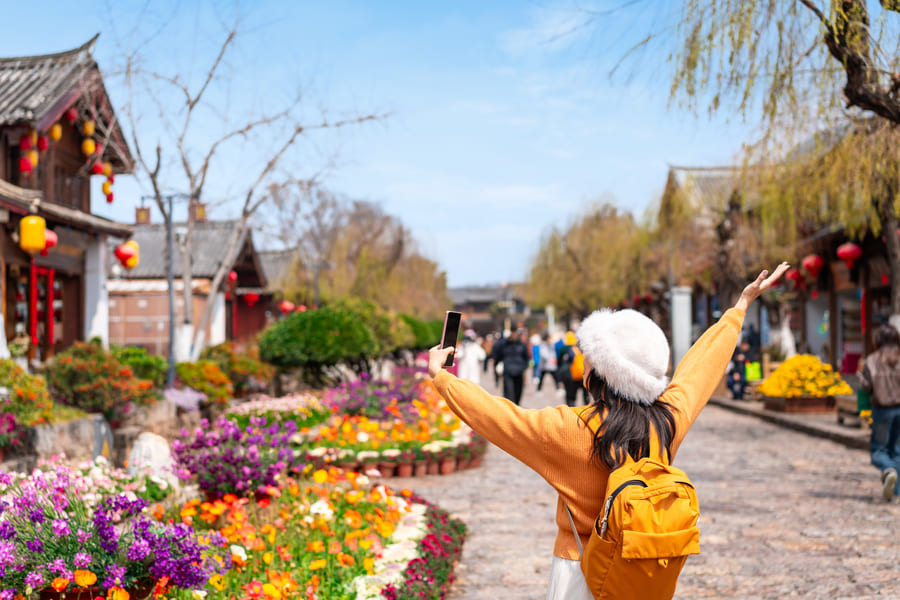 Turista con los brazos abiertos, sueter y mochila color naranja en las calles floreadas de Yunnan